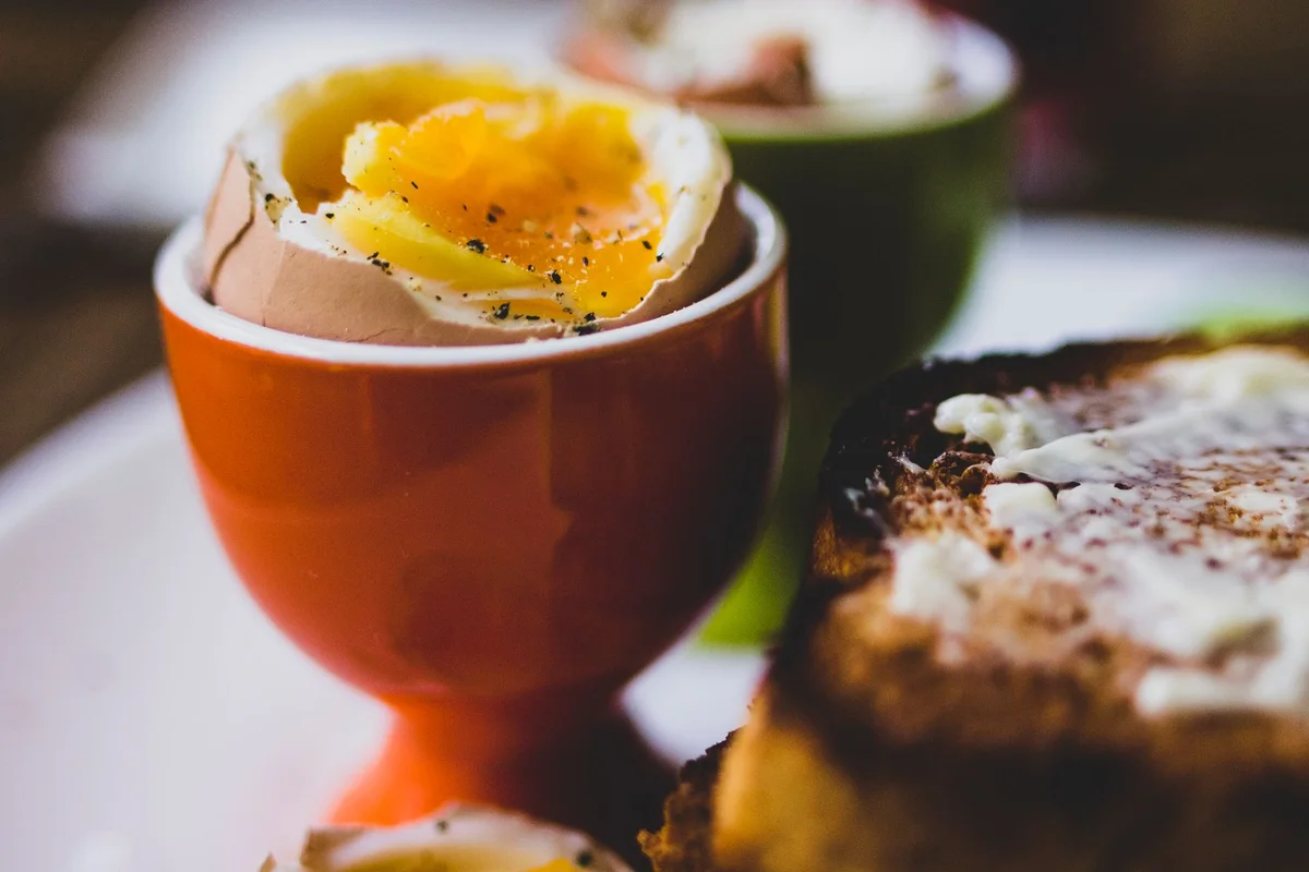 A soft-boiled egg in a ceramic cup beside buttered toast on a breakfast plate